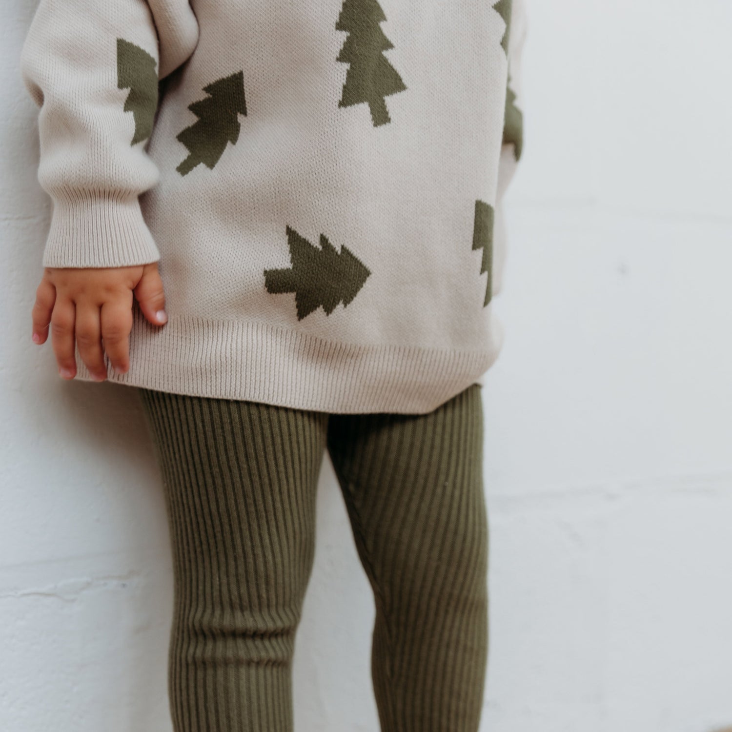 Child wearing a beige sweater with green tree patterns and green leggings against a white wall.